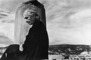 Artist Georgia O'Keeffe sitting draped in black cloak, looking downward with desert behind her