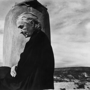 Artist Georgia O'Keeffe sitting draped in black cloak, looking downward with desert behind her