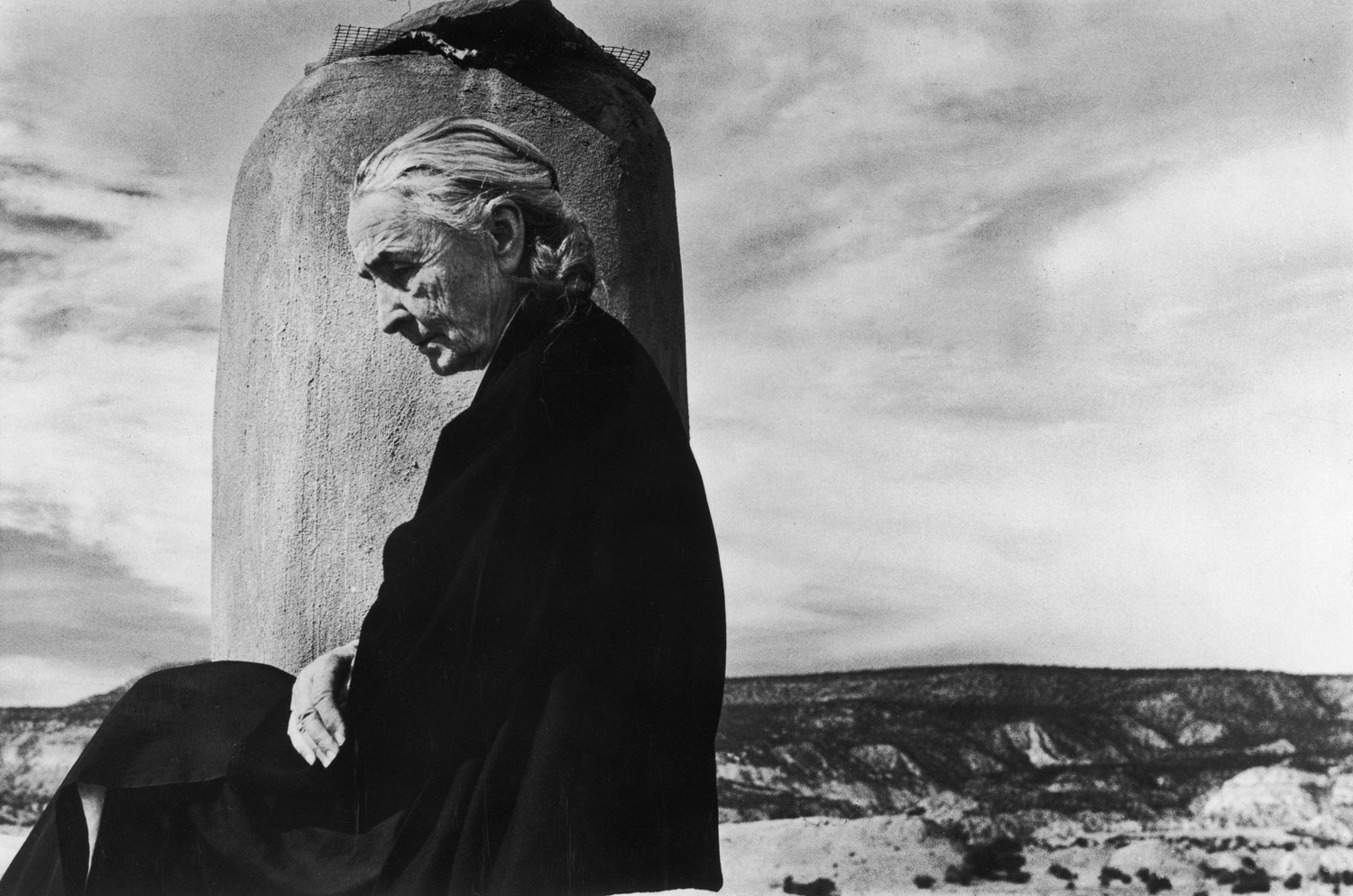 Artist Georgia O'Keeffe sitting draped in black cloak, looking downward with desert behind her